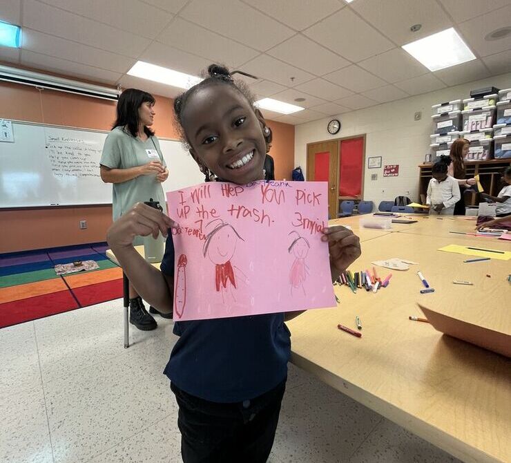 A student holds up her drawing on pink paper and smiles.