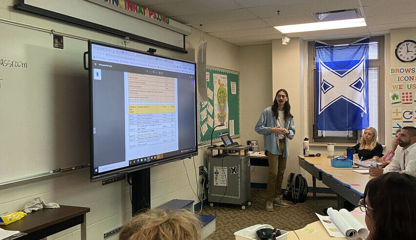 A man with long brown hair stands at the front of a classrooms presenting a document about Mindful Music Moments on a large screen.