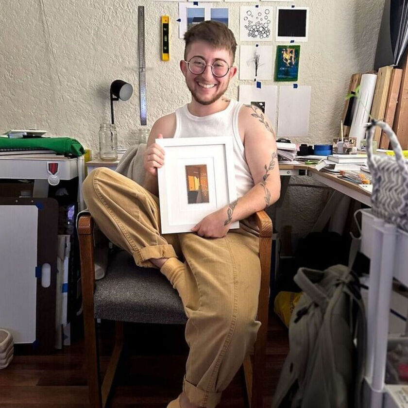 Elan smiles in a chair in an art studio holding a framed piece of art.