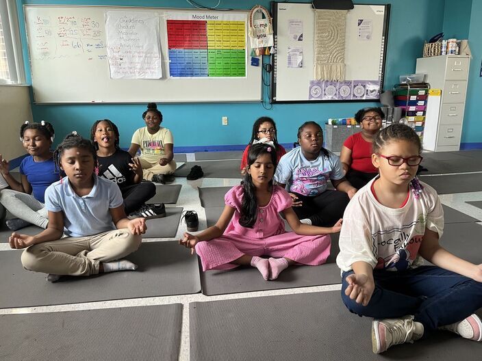 Nine students sit in meditative poses with their eyes closed on grey yoga mats.