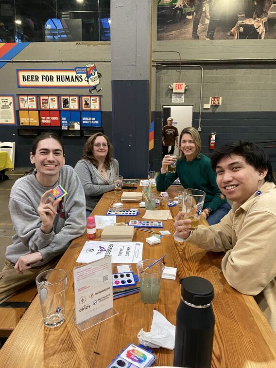 Four people sit at a table with paint, brushes, and beer.