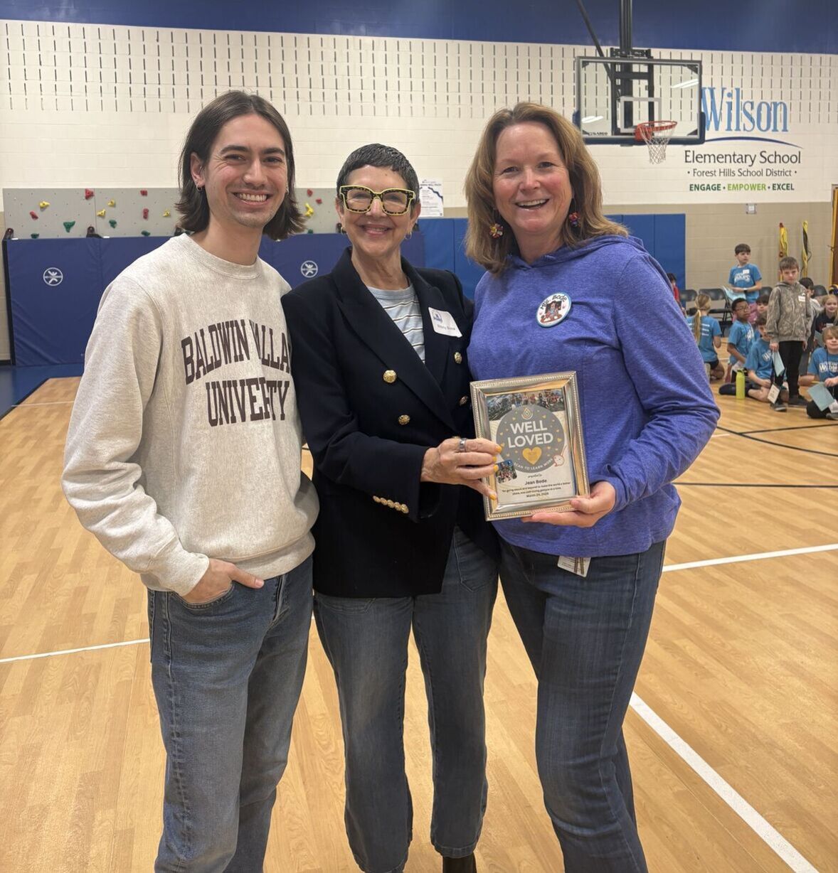Three people standing and smiling in a school gym. Jean is holding a picture frame that says Well Loved.
