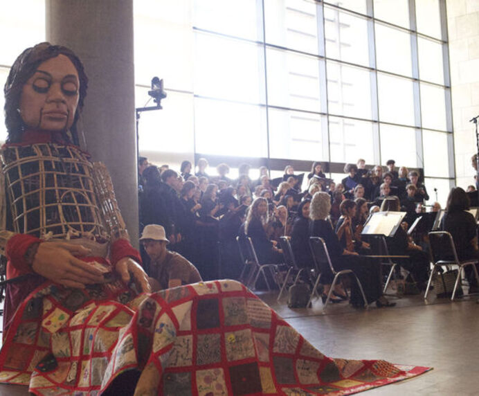 A 12-foot tall puppet sitting with a blanket over her legs. An orchestra plays in the background