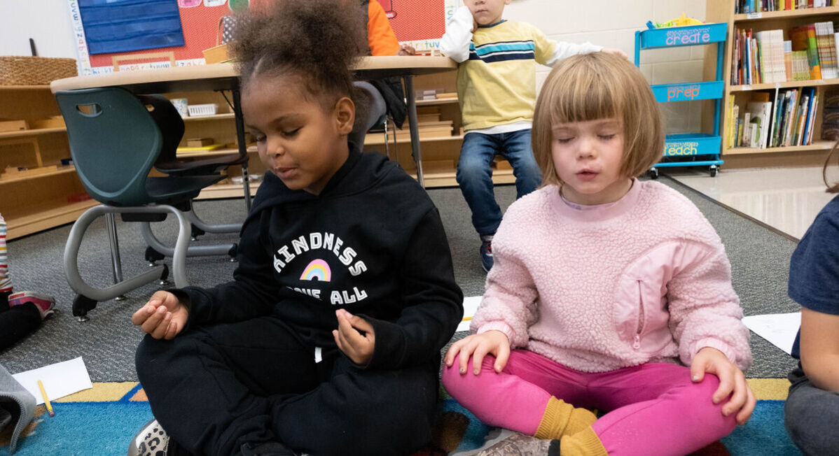 Two young students sit on a carpet with their legs crossed and their hands on their lap. Their eyes are closed as they take a deep breath.