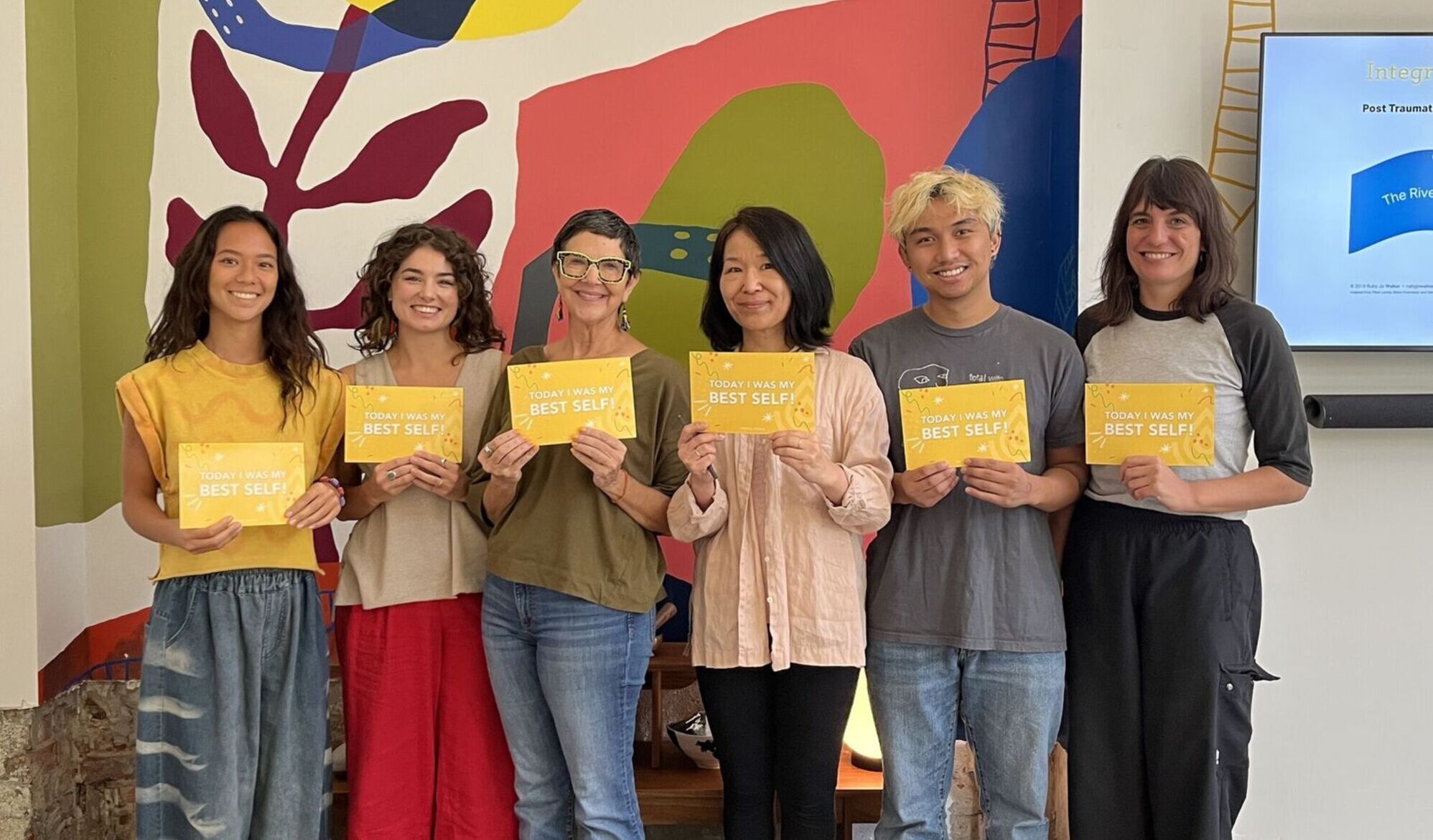 Six people stand in front of a colorful mural, holding yellow cards that say