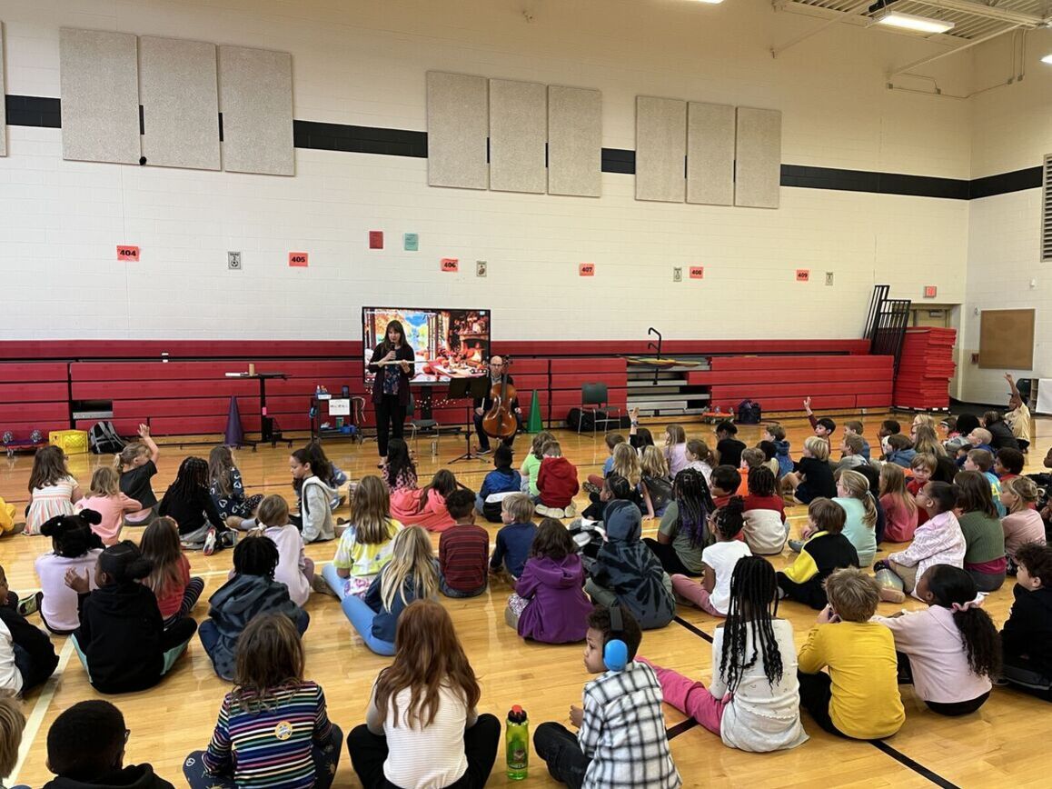 Students sit on the gym floor while Annie stands at the front with her flute.