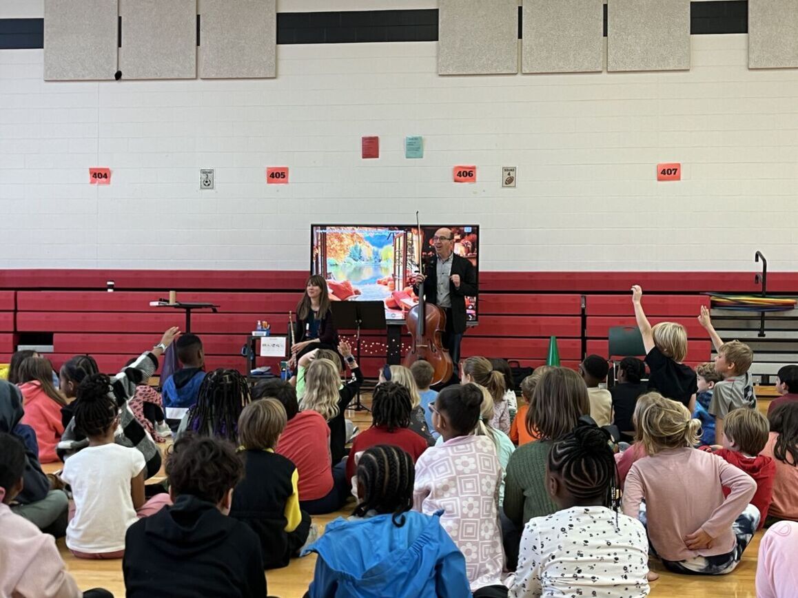 A group of students sits in the gym with their hands raised. Two musicians with a flute and cello stand in front of the group.