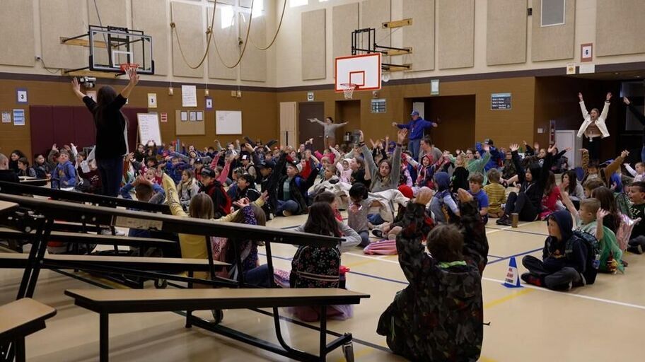 A gymnasium full of students sitting on the floor following their teachers in a breathing exercise by raising both arms slowly.