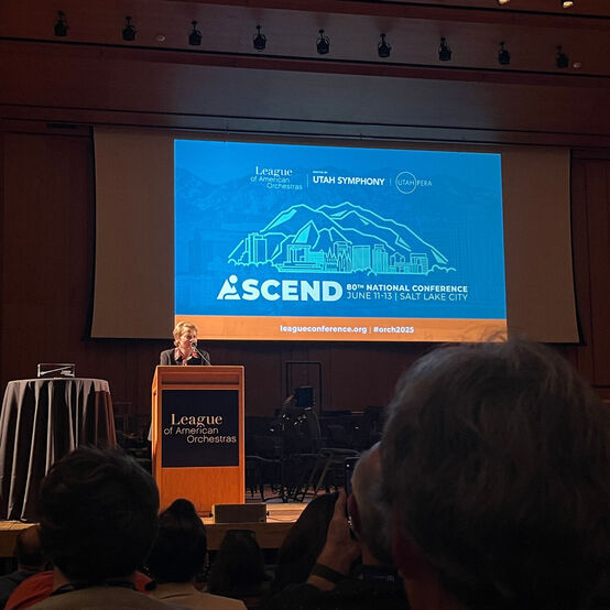 Marin Alsop speaking at a podium on a stage with the conference logo projected on a screen behind her.