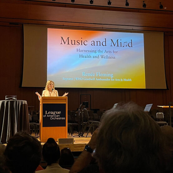 Renée Fleming speaking at a podium on a stage with ‘Music and Mind’ projected on a screen behind her.