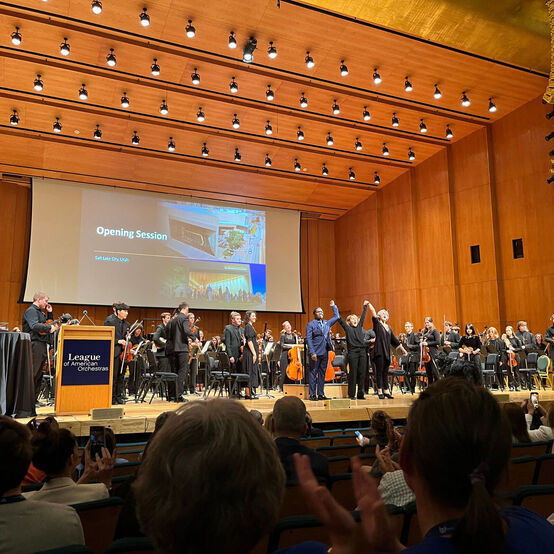 A conductor and two composers raise their hands to take a bow. A youth orchestra stands behind them while the audience applauds.