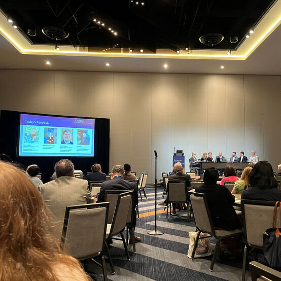 A panel of seven arts professionals sit on a stage in a hotel ballroom. Audience members sit in chairs at round tables.