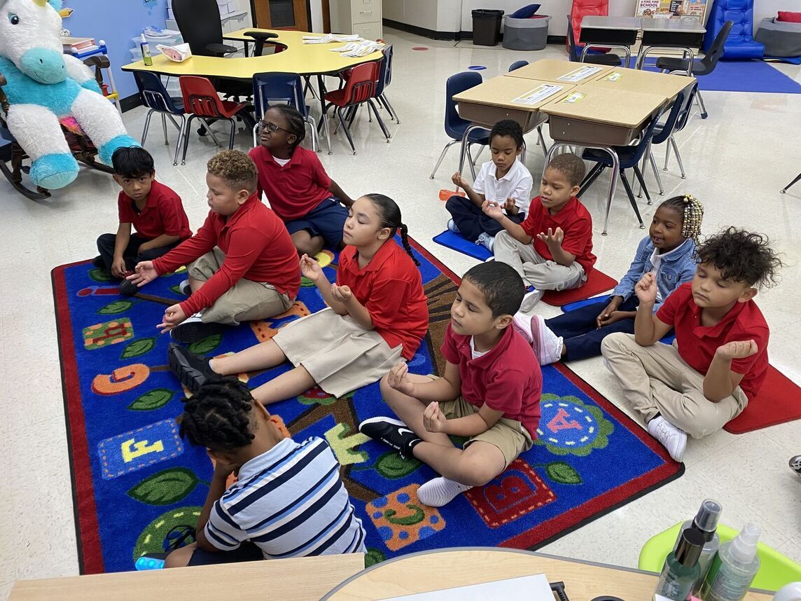 Ten students sit on a carpet in a classroom with their eyes closed and their hands in meditative positions.