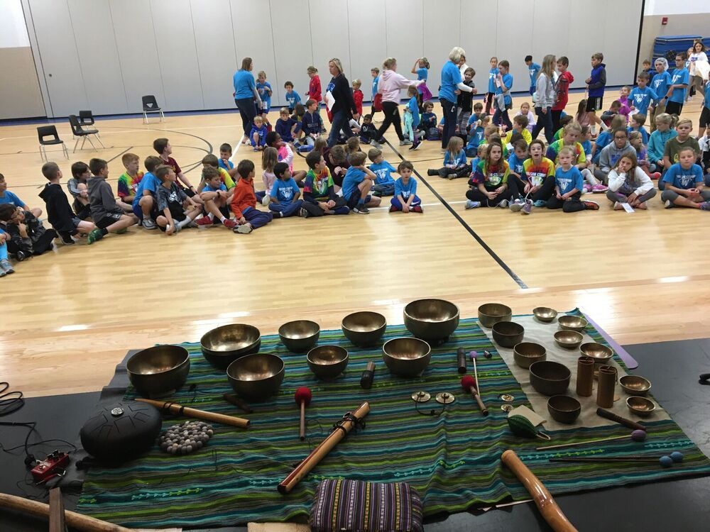 Forty students sit on a gymnasium floor with a large blanket of sound bowls and instruments in front of them.