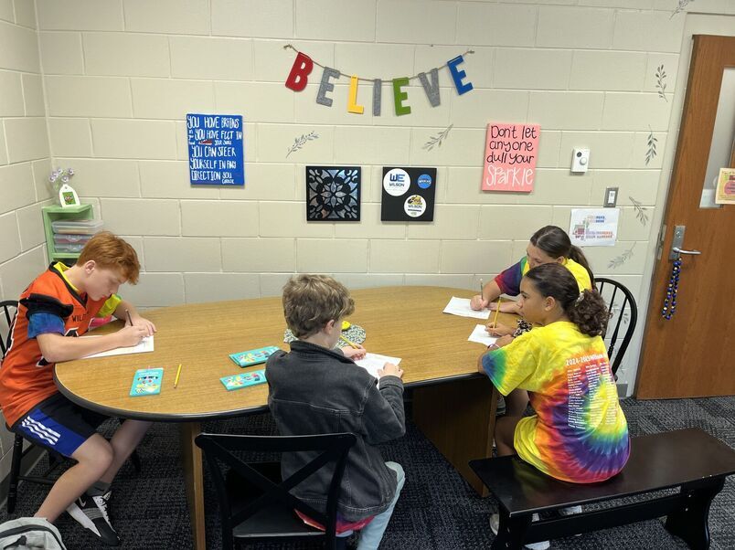 Four students sit around a table with colored pencils and worksheets.